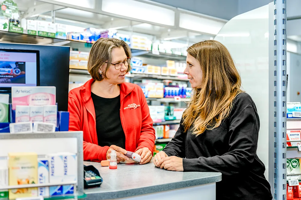 Ein Apotheker in einer roten Jacke spricht mit einem Kunden an der Apothekentheke über Medikamente. Im Hintergrund sind Regale mit verschiedenen Medikamenten und Gesundheitsprodukten zu sehen.