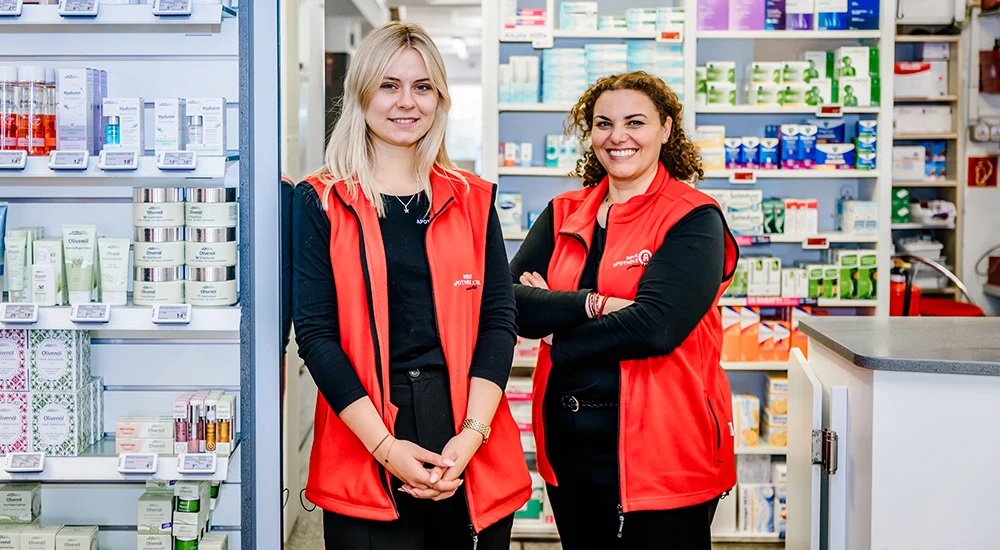 Zwei Frauen in roten Westen und schwarzen Hemden stehen lächelnd in einer Apotheke, umgeben von Regalen mit Gesundheits- und Schönheitsprodukten.
