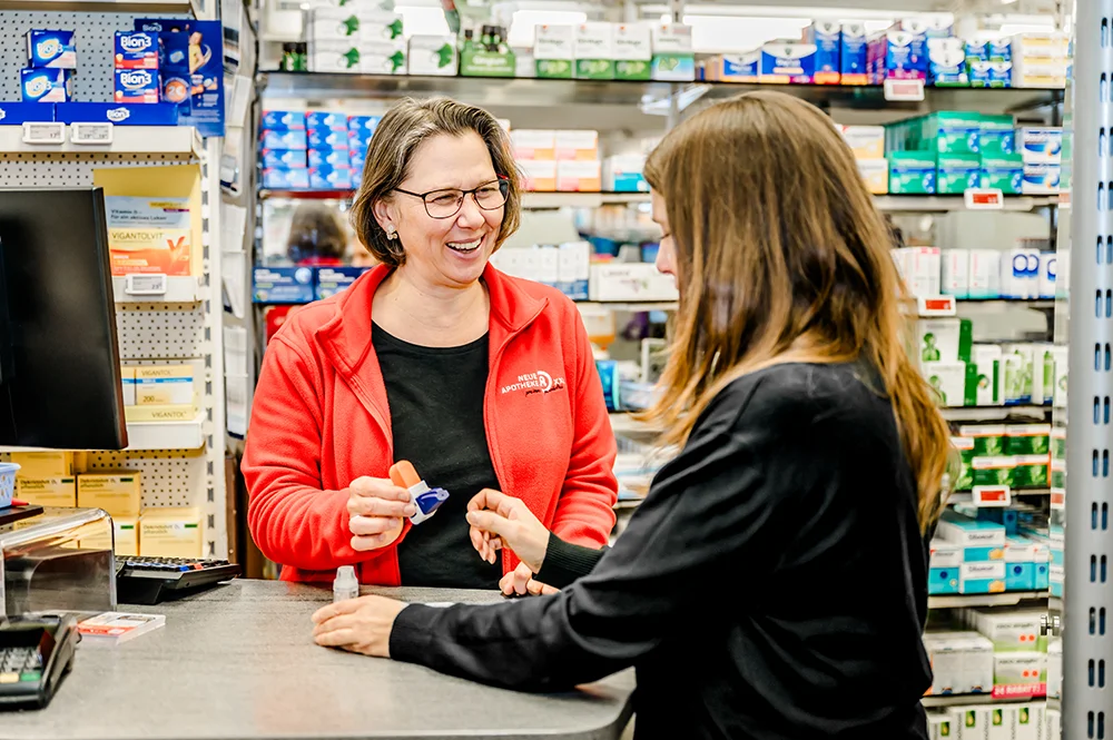 Ein lächelnder Apotheker in einer roten Jacke überreicht einem Kunden an einem Apothekentresen Medikamente, im Hintergrund stehen Regale mit Medikamenten und Gesundheitsprodukten.